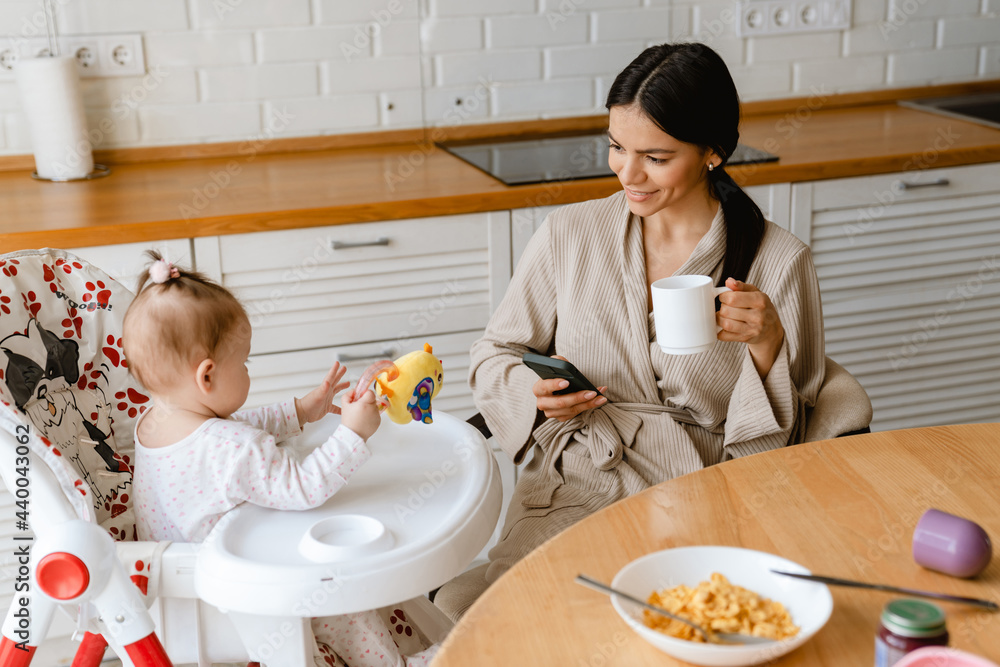 Young mother using mobile phone while having breakfast with her baby