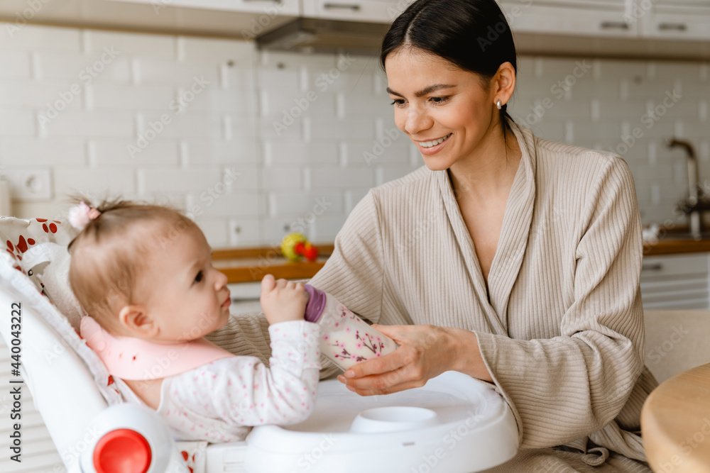 Young brunette mother smiling and feeding her baby in kitchen at home Young brunette mother smiling and feeding her baby in kitchen at home