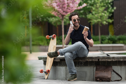 happy man in sunglasses holding paper cup and talking on smartphone near longboard and laptop. photo