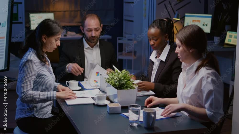 Diverse multi ethnic teamwork brainstorming company ideas analyzing financial strategy paperwork working overtime in office meeting room in evening. Coworkers discussing management presentation Diverse multi ethnic teamwork brainstorming company ideas analyzing financial strategy paperwork working overtime in office meeting room in evening. Coworkers discussing management presentation