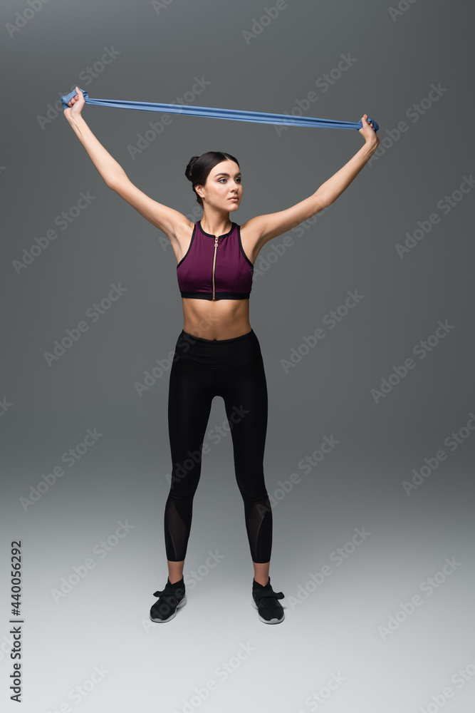 Obraz premium full length view of slim sportswoman training with resistance band on grey background.