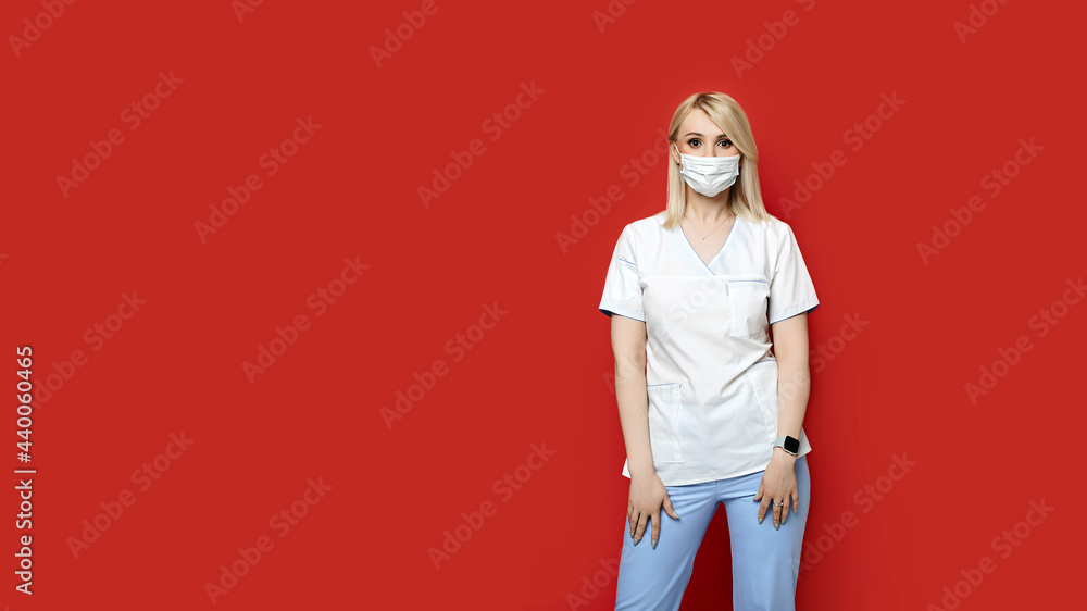 Confident female doctor wearing protective face mask standing on red background. Young woman intern in medical clothes posing.