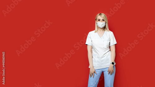 Obraz na płótnie Confident female doctor wearing protective face mask standing on red background. Young woman intern in medical clothes posing. w salonie