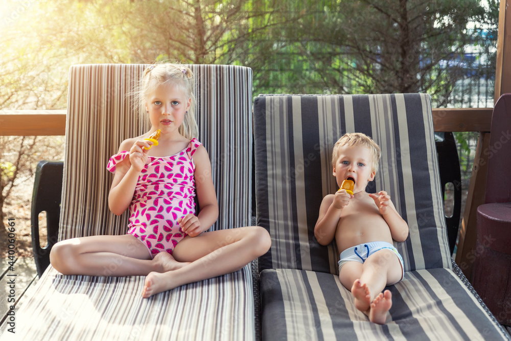 Fototapeta premium Two cute adorable caucasian blond little siblings enjoy having fun relaxing and eat fruit popsicle icecream sitting on sunbed in yard garden near pool sea beach. Brother and sister chilling outdoor