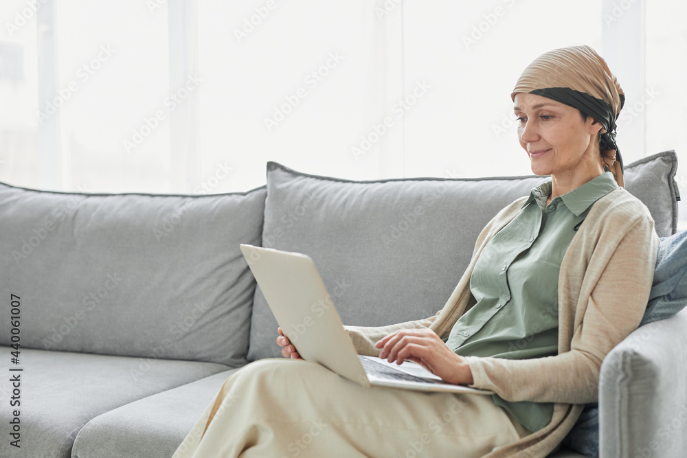 Candid portrait of mature woman using headscarf and using laptop while relaxing on couch in cozy home setting, copy space - obrazy, fototapety, plakaty 