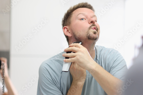 Young man shaving his beard with trimmer in front of mirror in bathroom