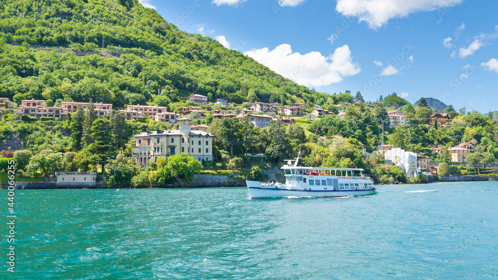 A ferry boat is leaving the village of Menaggio, Lake Como, Italy. Turquoise water on the foreground. Green hills with trees on the background.