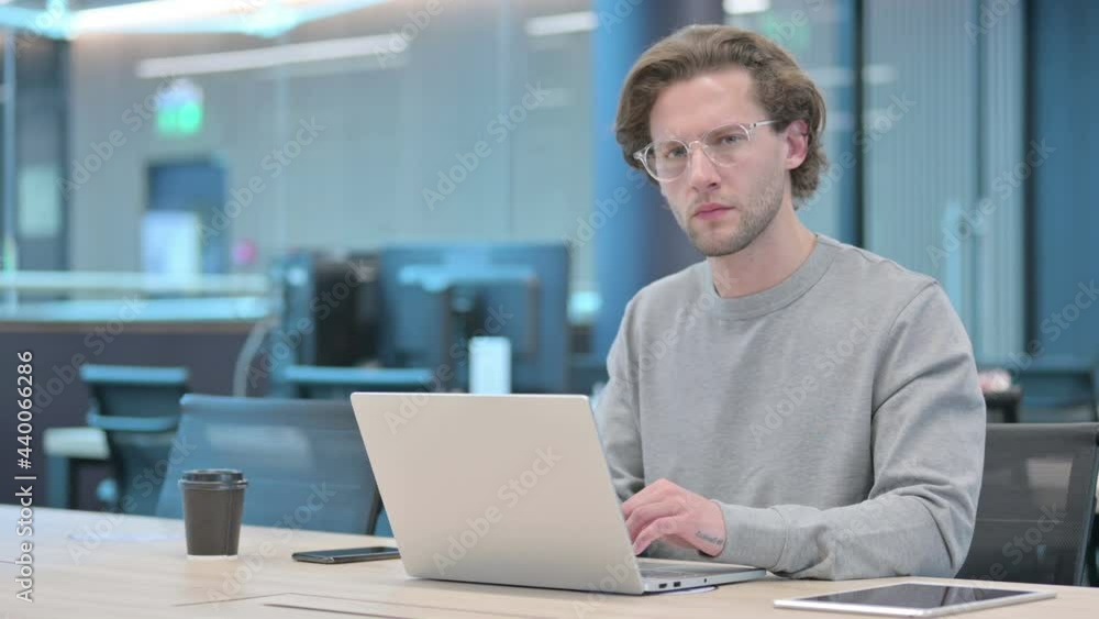 Young Businessman Looking at Camera while using Laptop in Office Young Businessman Looking at Camera while using Laptop in Office