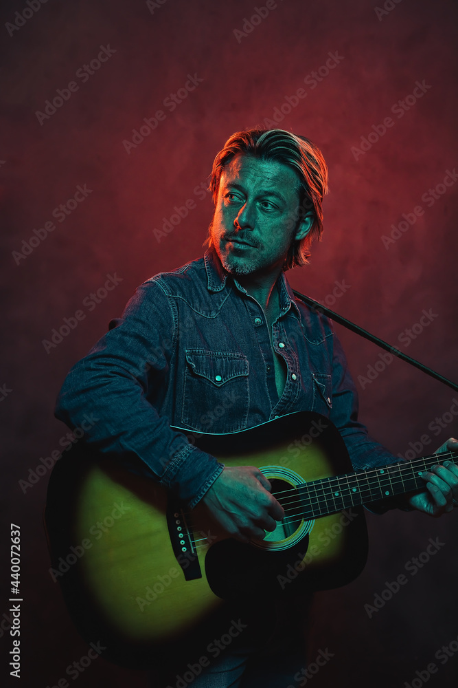 Man with acoustic western guitar in jeans shirt and jeans with belt in red and blue colored light.