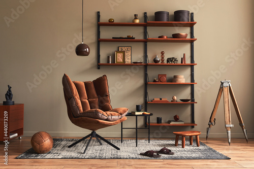 Stylish interior of living room with design brown armchair, wooden bookcase, pendant lamp, carpet decor, picture frames and elegant personal accessories in modern retro home decor. Template. photo