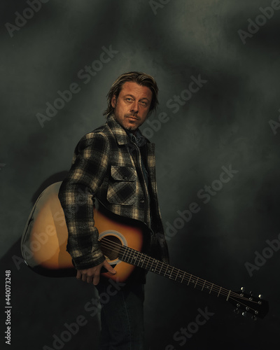 Fototapeta Naklejka Na Ścianę i Meble -  Man in checkered jacket with acoustic western guitar in dappled sunlight in front of a grey wall.