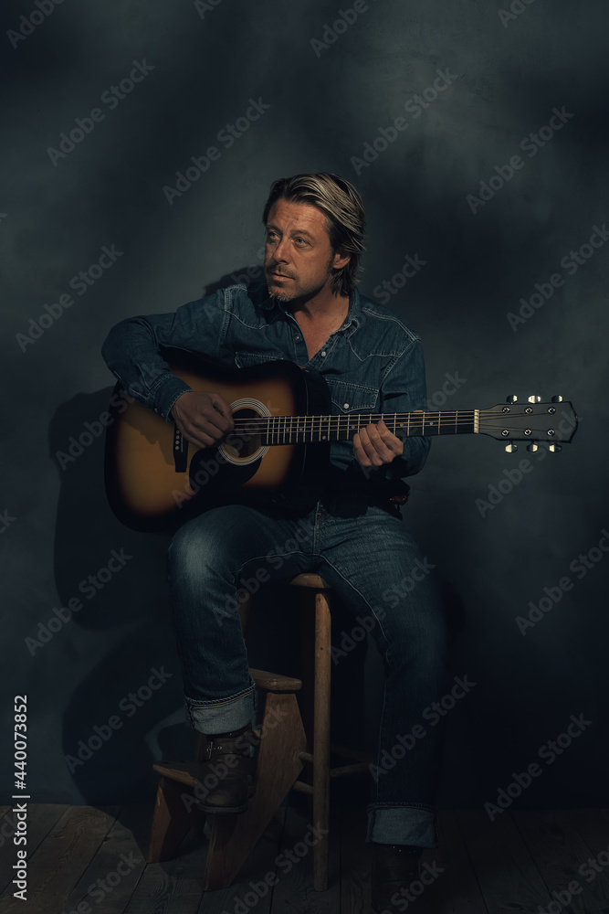 Obraz premium Man in denim shirt and jeans with acoustic western guitar on a wooden stool in dappled sunlight in front of a grey wall.