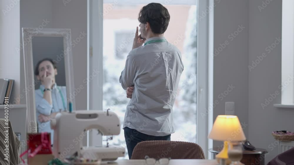 Back view of young skilled creative Caucasian male tailor looking out the window in atelier thinking touching mustache reflecting in mirror. Talented thoughtful professional man workplace in workshop Back view of young skilled creative Caucasian male tailor looking out the window in atelier thinking touching mustache reflecting in mirror. Talented thoughtful professional man workplace in workshop