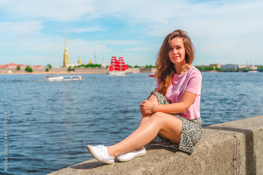 A beautiful young woman sits on the embankment on a summer day in St. Petersburg with a view of the Neva River and a ship with scarlet sails