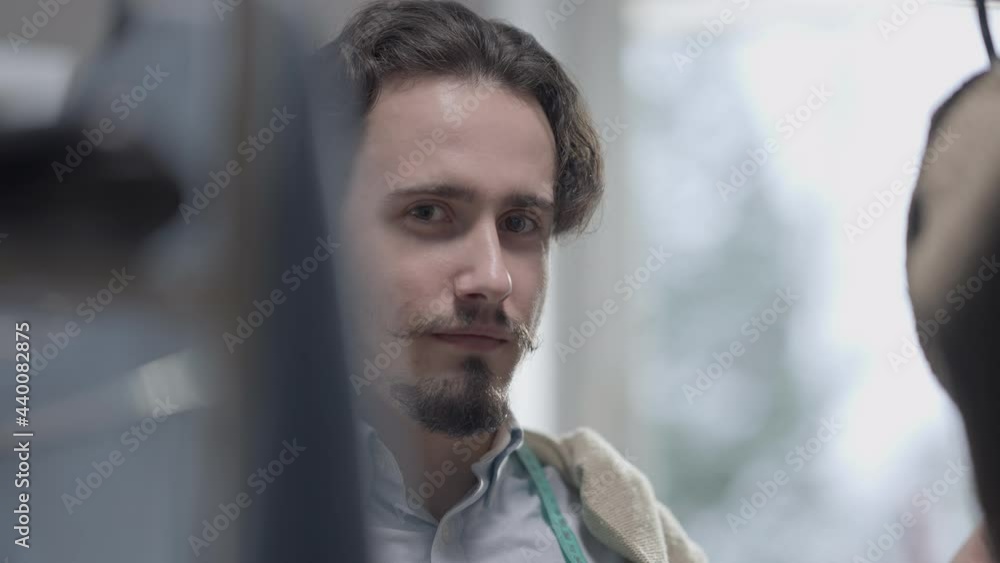 Close-up of positive skilled Caucasian male dressmaker looking through clothes on hangers looking at camera smiling. Talented confident man standing at rack in atelier posing. Fashion and skill Close-up of positive skilled Caucasian male dressmaker looking through clothes on hangers looking at camera smiling. Talented confident man standing at rack in atelier posing. Fashion and skill