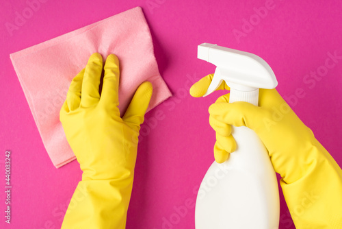 Top view photo of hands in yellow gloves holding pastel pink viscose rag and using detergent spray bottle without label on isolated pink background with copyspace
