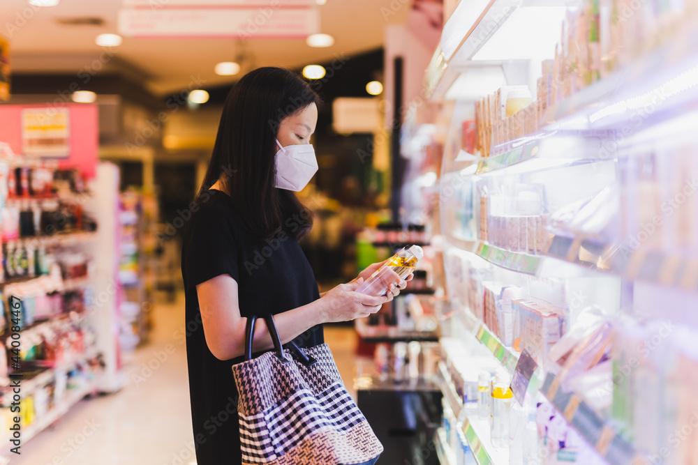 Asian woman in protective mask holding bottle of shampoo in supermarket.
