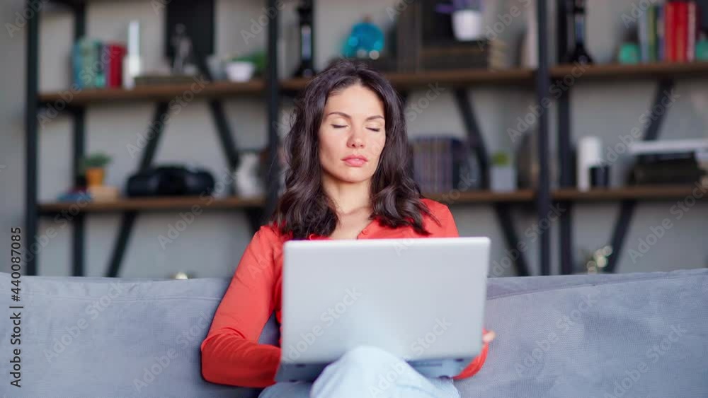 Front view of thoughtful middle aged woman typing on laptop computer sitting on sofa at home. Businesswoman working remotely from home and thinking Front view of thoughtful middle aged woman typing on laptop computer sitting on sofa at home. Businesswoman working remotely from home and thinking