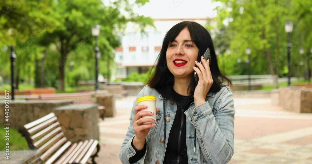 Brunette woman holding take away coffee and talking by phone, speaking by smartphone walking in park Brunette woman holding take away coffee and talking by phone, speaking by smartphone walking in park