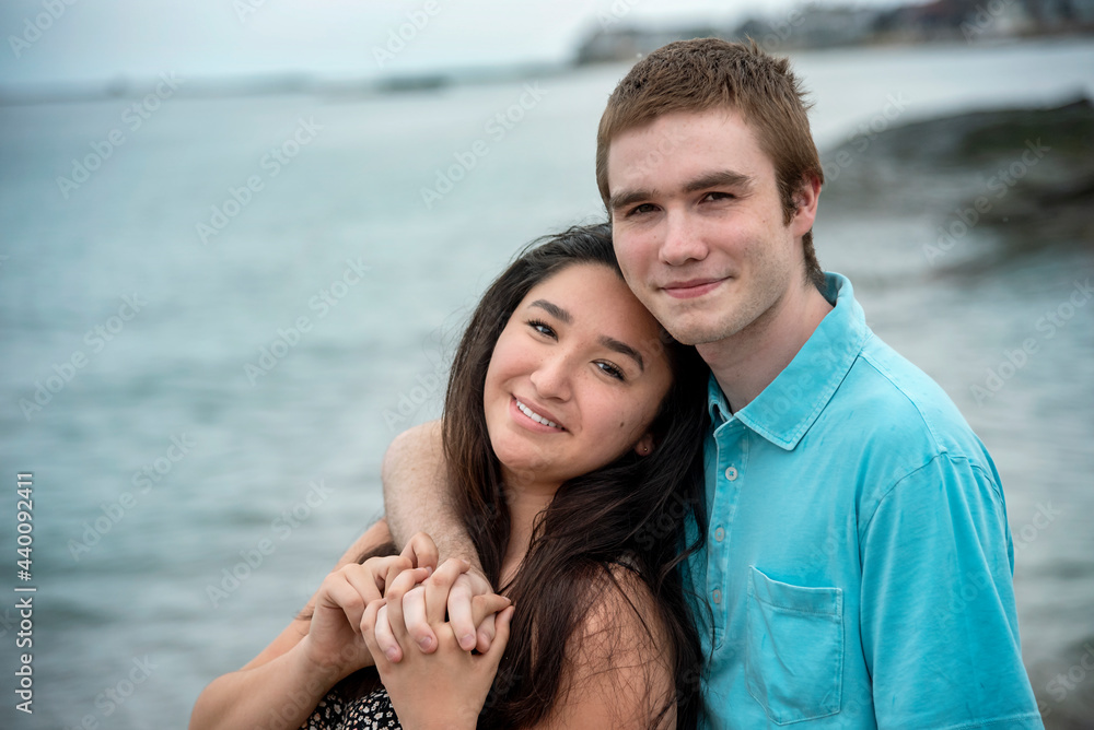 Portrait of young smiling happy couple in romantic casual loving embrace on a warm summer day at the beach with blue sky and water in background. Portrait of young smiling happy couple in romantic casual loving embrace on a warm summer day at the beach with blue sky and water in background.