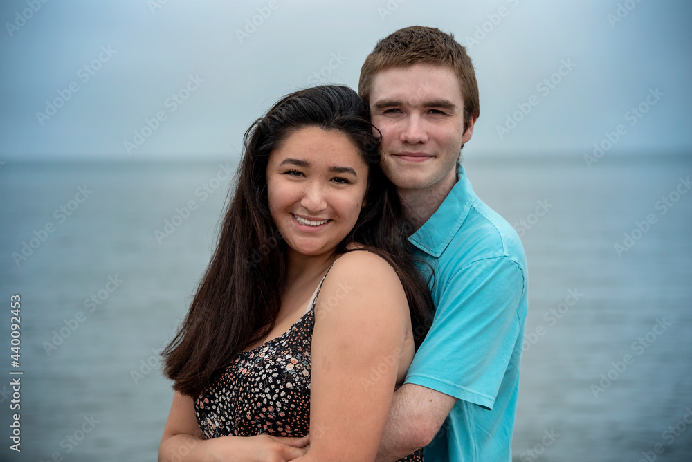 Young smiling happy couple in romantic casual loving embrace on a warm summer day at the beach with blue sky and water in background.