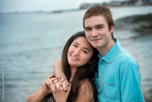Portrait of young smiling happy couple in romantic casual loving embrace on a warm summer day at the beach with blue sky and water in background.