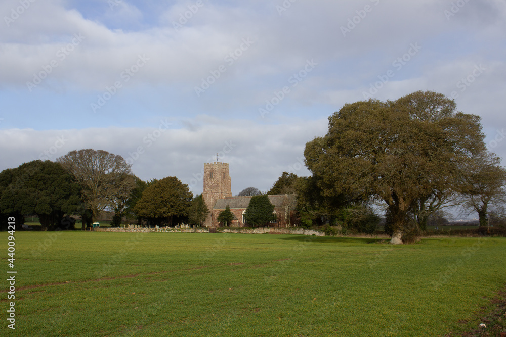 typical English village church surrounded by trees and farmland with a blue sky and white clouds