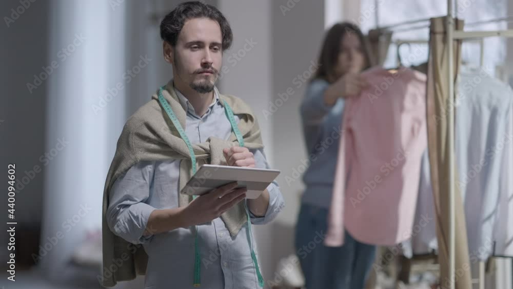 Young concentrated man surfing Internet on tablet as woman choosing clothes from new collection at background. Portrait of serious talented Caucasian fashion designer thinking on ideas in atelier Young concentrated man surfing Internet on tablet as woman choosing clothes from new collection at background. Portrait of serious talented Caucasian fashion designer thinking on ideas in atelier