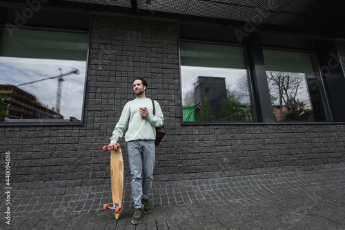 full length of man in sweatshirt holding paper cup and longboard outside.
