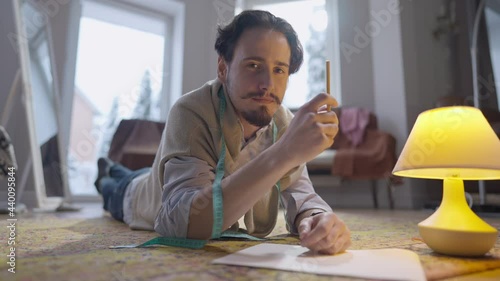 Young handsome man looking at camera lying on floor holding pencil. Portrait of talented creative Caucasian designer tailor posing in atelier designing collection sketches