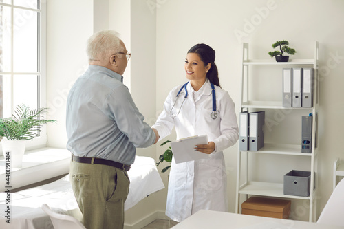 Friendly smiling woman physician meeting male elderly patient. Doctor and senior older man handshaking. Modern clinic office interior. Medicine, insurance and healthcare for retired people