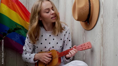 4k Young millennial hippie woman sitting on balcony play guitar. Music lesson and singer sitting. LGBTQ rainbow flag on background. Online music tuition. Playing instrument and enjoying songs 