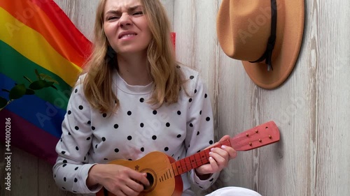 4k Young millennial hippie woman sitting on balcony play guitar. Music lesson and singer sitting. LGBTQ rainbow flag on background. Online music tuition. Playing instrument and enjoying songs 