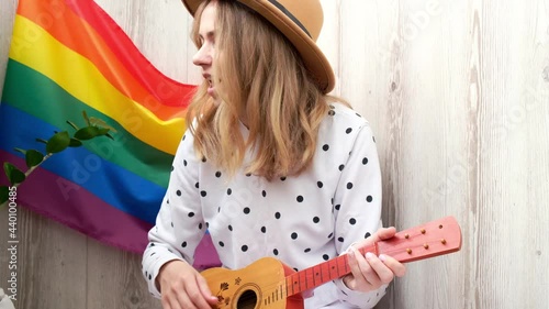 4k Young millennial hippie woman sitting on balcony play guitar. Music lesson and singer sitting. LGBTQ rainbow flag on background. Online music tuition. Playing instrument and enjoying songs 