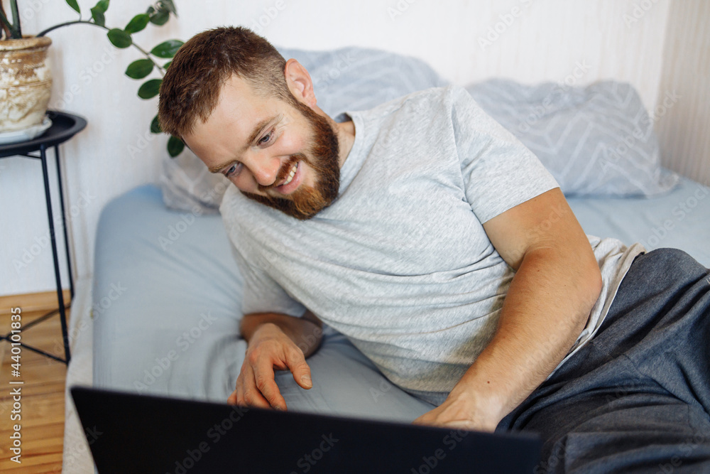 Close up portrait of European bearded man with red hair and big blue eyes sitting near laptop, concentrating and working on comfy sofa at home. Cozy interior with flora