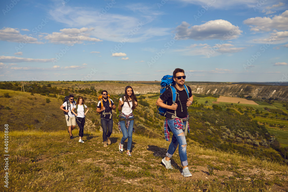 Hiking and traveling concept. Group of young friends hikers tourists traveling with backpacks on nature in row together with green valley landscape at background during summer vacations on sunny day Hiking and traveling concept. Group of young friends hikers tourists traveling with backpacks on nature in row together with green valley landscape at background during summer vacations on sunny day