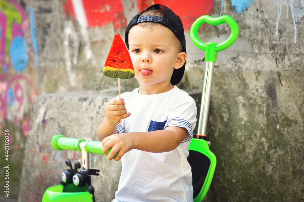 A cute toddler boy in street style clothes and a cap with a big watermelon candy in his hand sits on a scooter against a background of graffiti and licks sweet lips. A cute toddler boy in street style clothes and a cap with a big watermelon candy in his hand sits on a scooter against a background of graffiti and licks sweet lips.