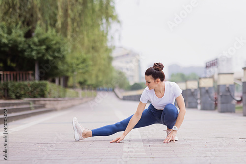 Runner stretching muscles outdoor at summer morning in city. Running athlete woman doing fitness stretch. Healthy young caucasian woman warming up outdoors before exercise.