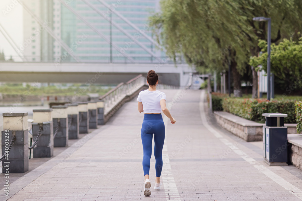 Young brunette Woman running on asphalt road in summer park. Active sporty caucasian female has morning workout. Athletic person in sportswear exercises outdoor. Dressed leggings and sportive top