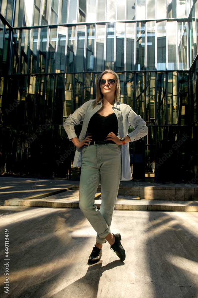 A young woman in a white shirt in light pants and sunglasses poses in the mirrored courtyard of the business center, illuminated by the sun's rays. Reflection in the mirror. Business portrait.