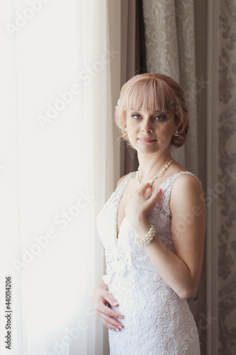 Pretty young Bride in elegant wedding dress near a window. Blonde-haired woman with wedding hair-style in royal room of hotel. Bride's morning in luxury interior. Wedding day ceremony