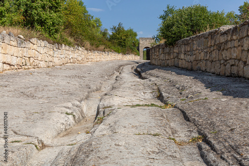 An ancient stone road with sagging ruts runs along the street along the stone fence to the arch .