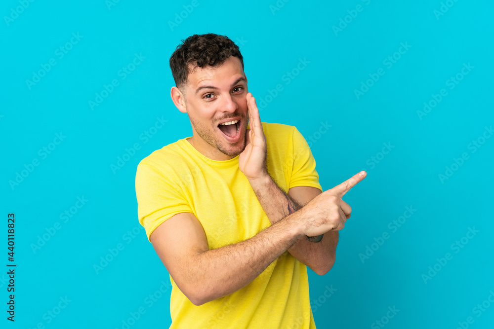 Young caucasian handsome man isolated on blue background pointing to the side to present a product and whispering something Young caucasian handsome man isolated on blue background pointing to the side to present a product and whispering something
