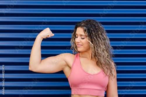 Sportswoman smiling while looking at bicep in front of corrugated wall photo