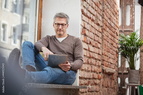 Mature man in eyeglasses using digital tablet while sitting on window sill at home photo