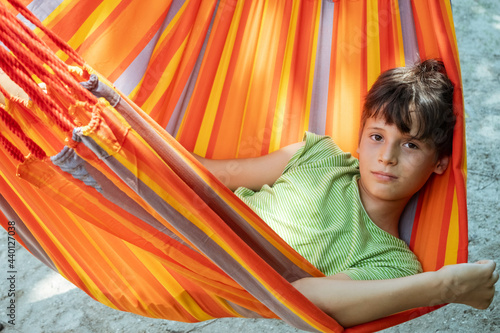 Caucasian teenager boy relaxing in bright striped orange hammock. Summer active leisure for kids. Child swinging on hammock. Teen on a colorful swing.