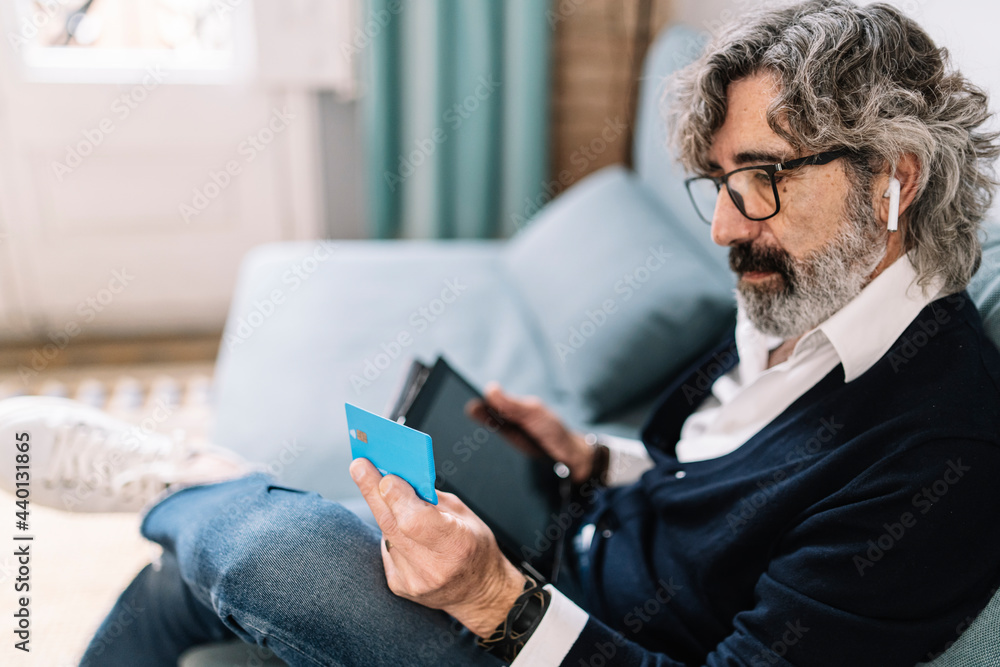 Handsome man looking at credit card holding digital tablet while sitting at home Handsome man looking at credit card holding digital tablet while sitting at home