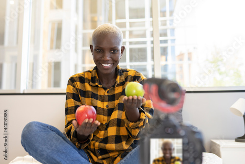 Smiling female influencer holding fruits while vlogging at home photo