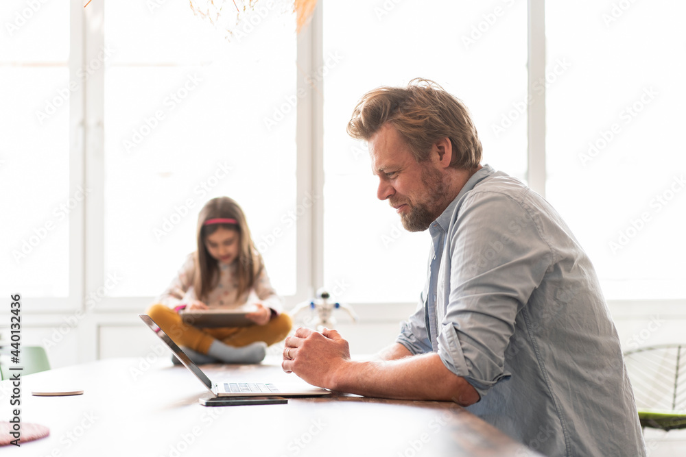 © Joseffson/Westend61 - Mature businessman working on laptop while girl sitting in background at home © Joseffson/Westend61 - Mature businessman working on laptop while girl sitting in background at home