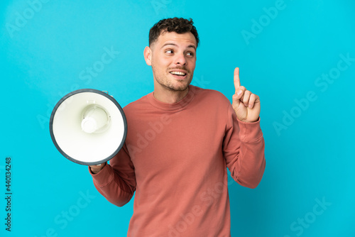 Young caucasian handsome man isolated on blue background holding a megaphone and intending to realizes the solution